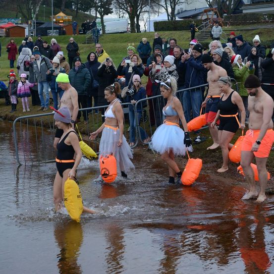 Eine Gruppe von Menschen watet in Badekleidung in einen Fluss, trägt Schwimmhilfen und orangefarbene Taschen, während eine Menge Zuschauer hinter einem Zaun steht.