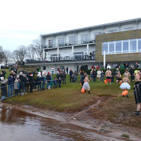 Eine Menschenmenge versammelt sich auf einem Rasenfeld und beobachtet mehrere Teilnehmer in Tüllröcken und Mützen, die auf einen Gewässerkörper zulaufen. Ein Gebäude mit Balkonen und Fenstern ist im Hintergrund.
