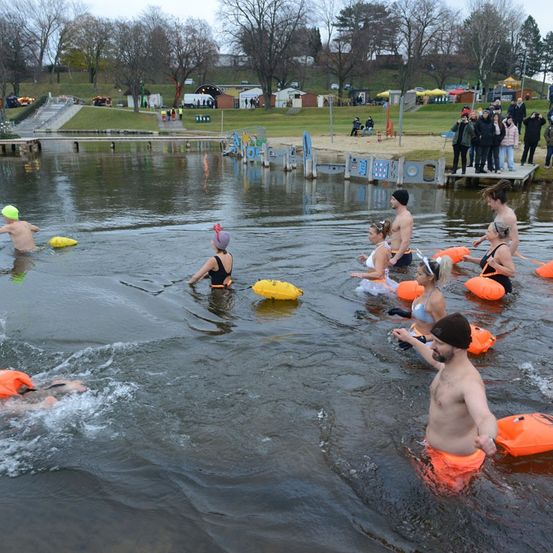 Teilnehmer eines Winterschwimmevents, mit Schwimmmützen und Schwimmwesten, steigen ins kalte Wasser, während andere vom Ufer aus beobachten.