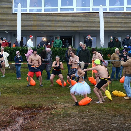 Eine Gruppe von Menschen in Badekleidung, einige mit Weihnachtsmützen, spielen mit orangefarbenen Luftballons auf einem Feld. Zuschauer beobachten hinter einer Barriere, und ein Gebäude mit Glasfenstern ist im Hintergrund.