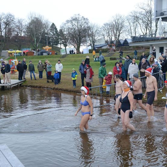 Eine Gruppe von Menschen in Badekleidung und Mützen geht in einem Fluss. Zuschauer beobachten vom Flussufer aus, einige tragen Weihnachtsmützen. Die Szene ist von einem Grasbereich mit Bäumen und Gebäuden umgeben.