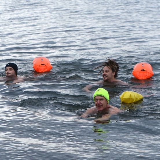 Drei Männer schwimmen in einem Gewässer, jeder mit einem Rettungsboje für die Sicherheit. Einer trägt eine grüne Mütze, ein anderer eine schwarze Mütze, und der dritte hat einen Irokesenschnitt.
