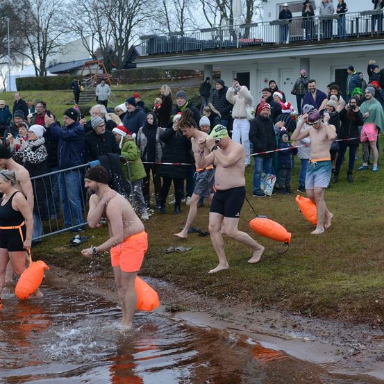 Eine Gruppe von Menschen in orangefarbenen Shorts betritt einen Fluss. Zuschauer beobachten hinter einem Seilbarriere. Einige haben Kameras, und einige tragen Mützen. Bäume und ein Gebäude sind im Hintergrund.