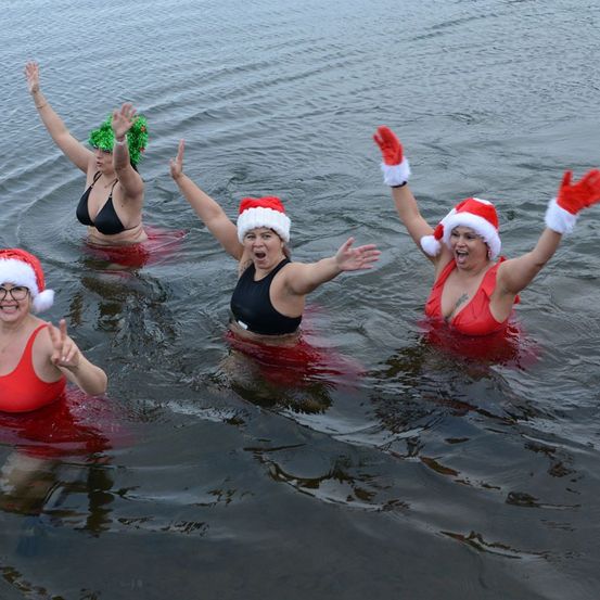 Vier Frauen sind im Wasser und tragen Weihnachtsmützen und rote Badeanzüge. Sie haben ihre Hände erhoben und scheinen Spaß zu haben. Das Wasser ist ruhig.