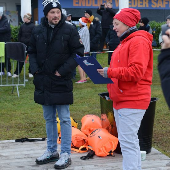 Ein Mann in einem schwarzen Mantel steht mit einem Klemmbrett, während eine Frau in einem roten Mantel eine blaue Mappe in der Hand hält. Sie sind umgeben von Rettungswesten und einem Mülleimer auf einem Rasenfeld mit Zuschauern im Hintergrund.
