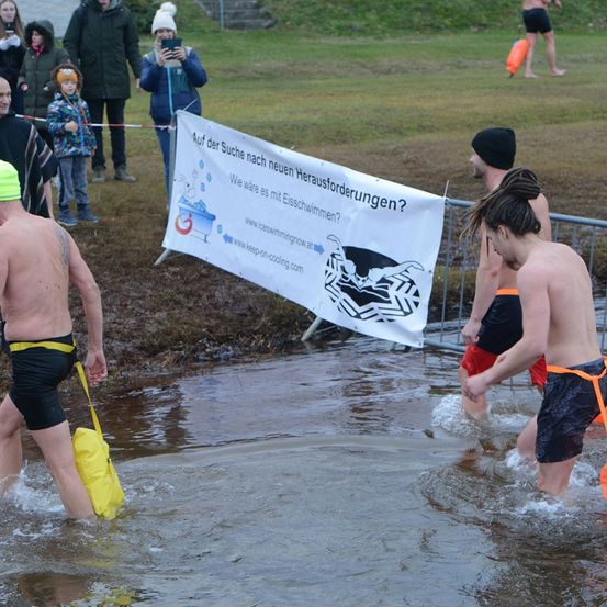 Eine Gruppe von Männern geht in ein Gewässer, an dem ein Banner für Eisschwimmen wirbt. Zuschauer beobachten von den Seiten.
