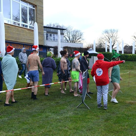 Eine Gruppe von Menschen in Badekleidung und Weihnachtsmützen steht hinter einem Hindernis in einer Reihe, möglicherweise in einem Park auf ein Ereignis wartend.