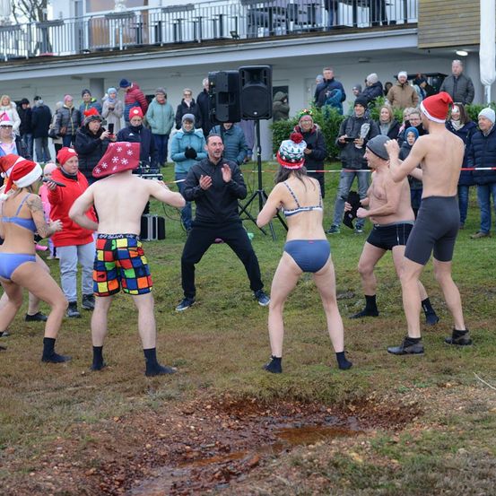 Eine Menschenmenge beobachtet eine Gruppe von Menschen, die auf einem Grasfeld tanzen, einige in Badekleidung, andere mit festlichen Mützen. Ein Mann in einem schwarzen Mantel klatscht.