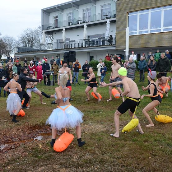 Eine Gruppe von Menschen nimmt an einer festlichen Veranstaltung teil, trägt Tüllröcke und hält orangefarbene Taschen, während eine Menge hinter einer Absperrung zuschaut. Ein Gebäude mit Balkonen und Glasfenstern ist im Hintergrund.