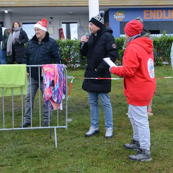 Ein Mann in einem schwarzen Mantel und einer Weihnachtsmütze spricht in ein Mikrofon. Ein anderer Mann in einem roten Pullover hört zu und hält ein Papier. Dahinter ist ein Zaun mit bunten Tüchern behängt.