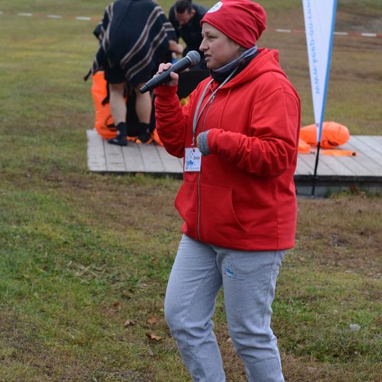 Eine Frau in einem roten Hoodie steht auf einer Holzplattform, hält ein Mikrofon und spricht. Hinter ihr knien sich zwei Personen, und ein orangefarbener Beutel liegt auf dem Boden. Der Hintergrund zeigt ein saftiges Feld mit einem weißen Banner.