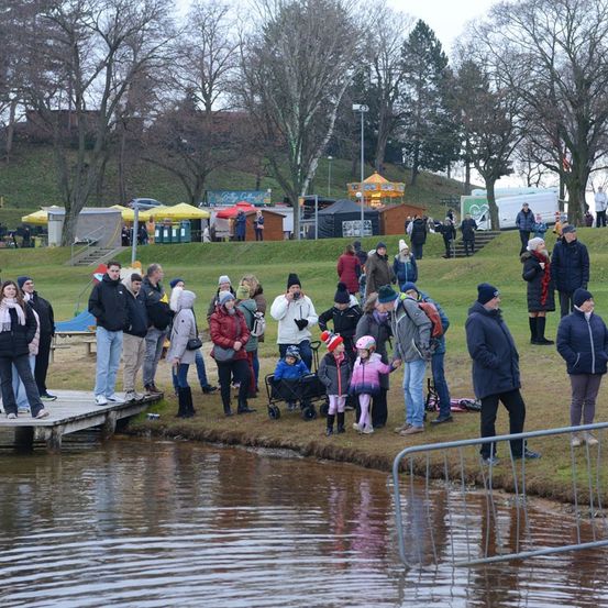 Eine Menschenmenge steht in der Nähe eines Sees, einige in Winterkleidung. Einige sind auf einem Steg, andere auf dem Gras, mit Zelten und einem Karussell im Hintergrund.