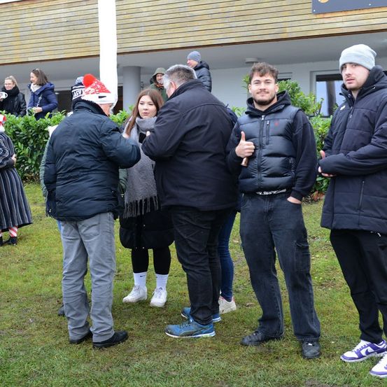 Mehrere Personen stehen draußen in der Nähe eines Gebäudes mit einem Busch und einer Säule. Ein Mann gibt einen Daumen hoch, während andere sprechen.