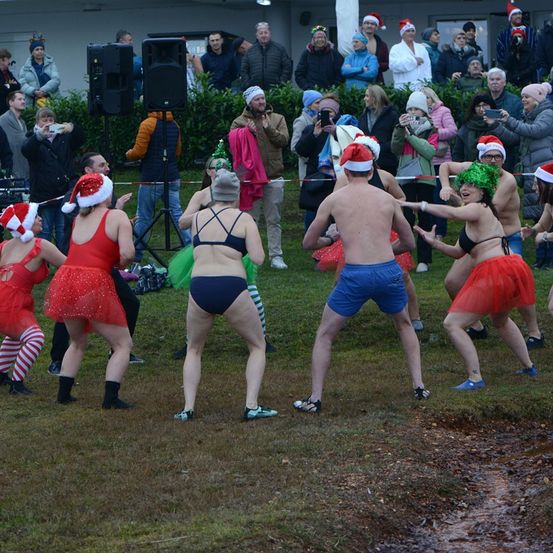 Teilnehmer einer festlichen Veranstaltung, die Weihnachtsmützen und Badeanzüge tragen, tanzen auf einem Rasenfeld, während Zuschauer hinter einer Seilbarriere zusehen.