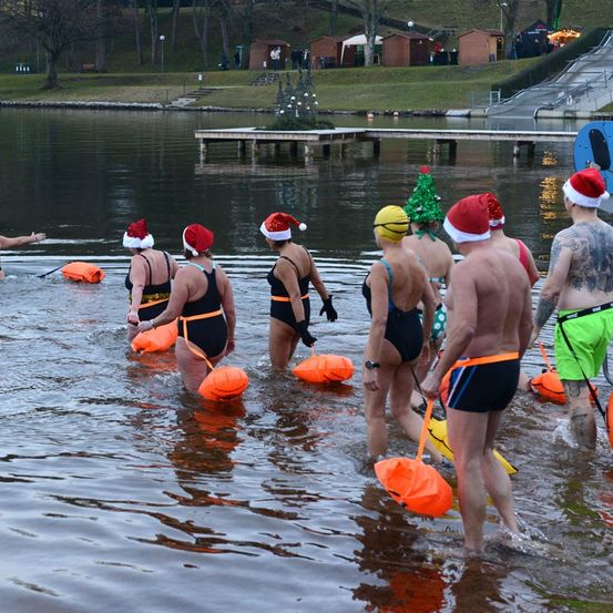 Eine Gruppe von Menschen in Badekleidung, die Weihnachtsmützen tragen, watet in einen See, jeder trägt orangefarbene Schwimmhilfen. Im Hintergrund steht ein festlicher Baum und mehrere kleine Häuser.