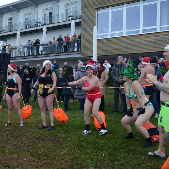 Eine Gruppe von Menschen in Badekleidung und Weihnachtsmützen nimmt an einem Rennen auf einem Rasenfeld teil. Sie tragen orangefarbene Taschen, und andere beobachten von einem Gebäude mit Balkonen aus.