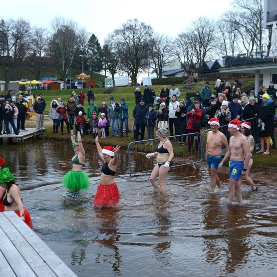 Eine Gruppe von Menschen in Badekleidung und festlichen Mützen watet während einer Winterveranstaltung in einem See, während eine Menge vom Ufer aus zusieht.