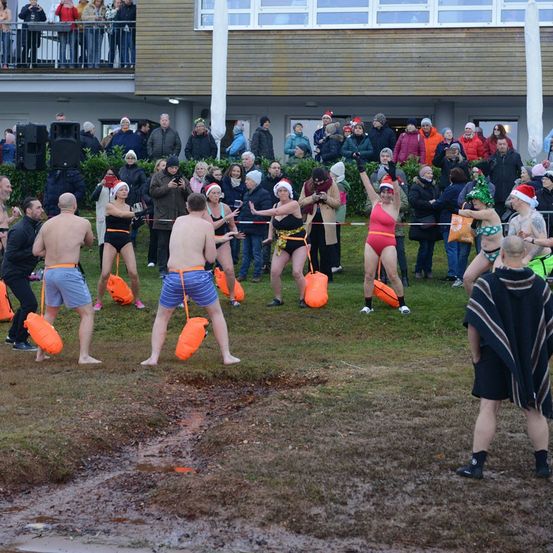 Eine Gruppe von Menschen nimmt an einem Winterschwimmen vor einer Menge teil, die festliche Mützen und Mäntel trägt. Einige Schwimmer sind in Badeanzügen und halten orange Taschen, bereit zum Sprung ins Wasser.