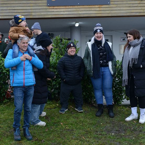 Eine Gruppe von Menschen steht vor einem Gebäude, lächelt und posiert für ein Foto. Sie tragen alle Winterkleidung. Ein Mann hält ein Kind auf seinen Schultern.