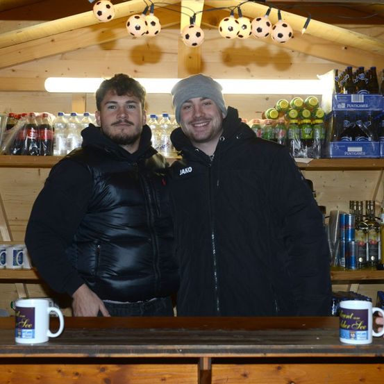 Zwei Männer in schwarzen Jacken posieren in einem Holzschuppen, mit Flaschen und Fußballen darüber. Tassen stehen auf dem Tresen vor ihnen.