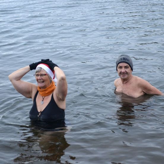 Zwei Personen im Wasser, eine trägt einen Weihnachtshut und Handschuhe, die andere eine Mütze. Sie lächeln beide.