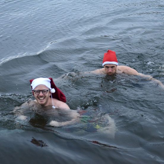 Zwei Männer schwimmen im Meer, einer trägt einen Weihnachtshut und eine Brille, der andere einen roten Weihnachtshut, beide lächeln im kalten Wasser.