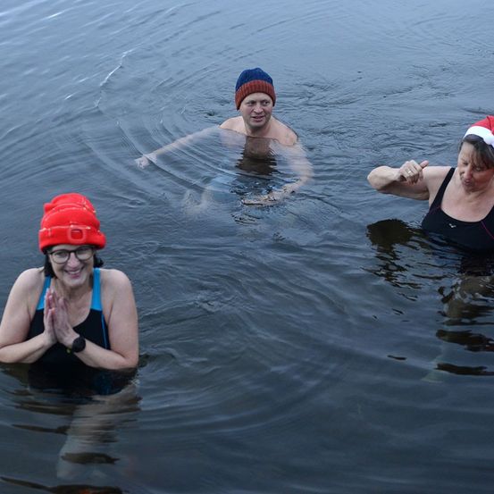 Drei Personen in Badekleidung, darunter ein Mann in der Mitte und zwei Frauen an den Seiten, befinden sich in kaltem Wasser und nehmen wahrscheinlich an einem Winterschwimmen teil.