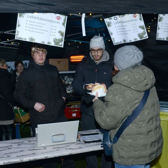 Eine Gruppe von Menschen steht bei Nacht vor einem Zelt, mit einem Tisch, auf dem ein Laptop und Papiere liegen. Ein Mann gibt einer Frau ein Sandwich. Menütafeln hängen oben, und Menschen versammeln sich in der Nähe.