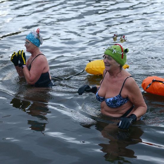 Zwei Frauen in Badeanzügen und Wintermützen stehen im Wasser und tragen Handschuhe. Eine hat eine Taucherbrille und eine blaue Kopfband mit einer Feder, die andere hat eine grüne Mütze mit Rehenohren.