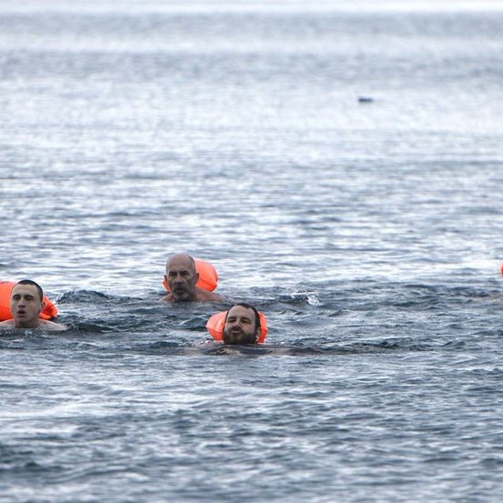 Drei Männer mit orangefarbenen Schwimmwesten schwimmen im ruhigen Ozean. Der Mann in der Mitte hat einen Bart und einen Schnurrbart.