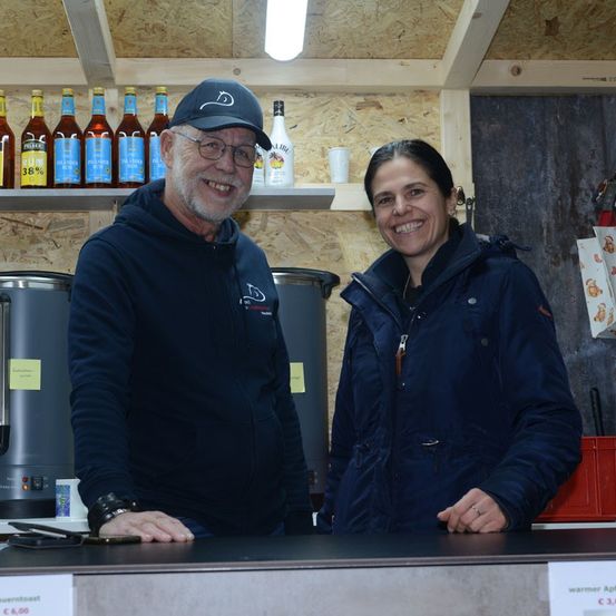 Zwei Menschen stehen lächelnd hinter einem Tresen. Dahinter befinden sich Regale mit Flaschen und Tassen. Einer trägt eine blaue Kapuzenjacke und der andere eine blaue Jacke. Es gibt auch zwei große graue Behälter.