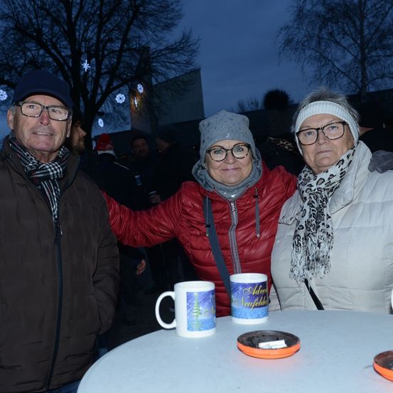 Drei Personen posieren nachts draußen für ein Foto. Sie haben Tassen auf einem Tisch vor sich. Die Frau in der Mitte trägt eine rote Jacke. Sie sind alle warm für das kalte Wetter gekleidet.