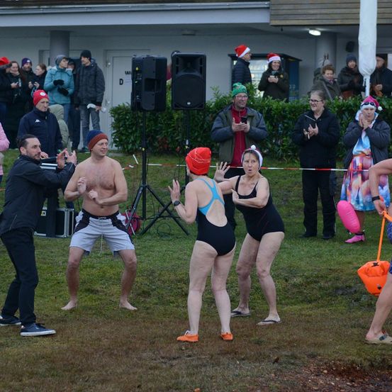 Eine Gruppe von Menschen, einige in Badekleidung, führt einen Tanz auf einem Rasenfeld mit einem Gebäude im Hintergrund auf. Zuschauer beobachten sie.