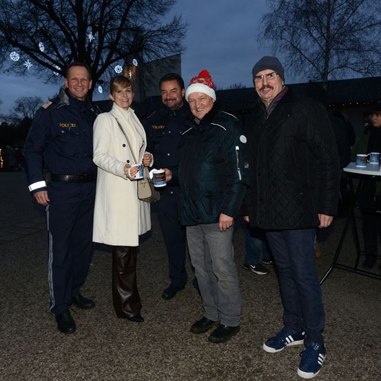 Fünf Personen stehen bei Nacht draußen, möglicherweise bei einem Feiertagsereignis. Einer hält Tassen, und alle tragen Winterkleidung. Bäume mit Ornamenten stehen hinter ihnen.