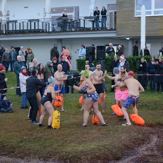 Eine Gruppe von Menschen, die in Badekleidung und Mützen gekleidet sind, versammelt sich draußen in der Nähe eines Gebäudes. Sie halten orangefarbene Schwimmhilfen und scheinen sich auf eine Veranstaltung vorzubereiten. Einige jubeln, während andere herumstehen.