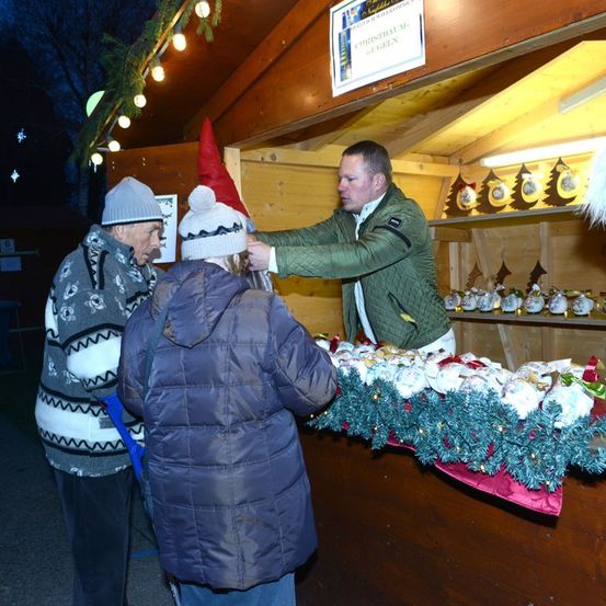 Drei Personen versammeln sich bei Nacht an einem Weihnachtsmarktstand. Ein Mann in einer grünen Jacke passt die Mütze eines Kindes an, während ein älterer Mann in einer Mütze neben ihnen steht.