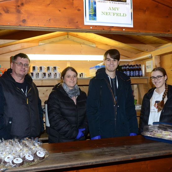 Vier Personen stehen in einem hölzernen Kiosk mit einem Schild über ihnen, das AMV Neufeld steht. Es gibt Tassen und Flaschen an den Regalen. Vor ihnen steht ein hölzerner Tresen mit verschiedenen Desserts.