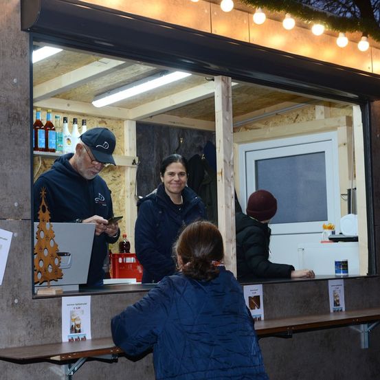 Eine winterliche Szene mit einem kleinen Laden. Drinnen bedient ein Mann in einer Kappe die Kasse, während ein anderer Mann neben ihm steht. Eine Frau und eine weitere Person stehen vor dem Laden und bestellen Getränke. Der Laden ist mit warmen Lichtern beleuchtet und mit Weihnachtsdekoration geschmückt.