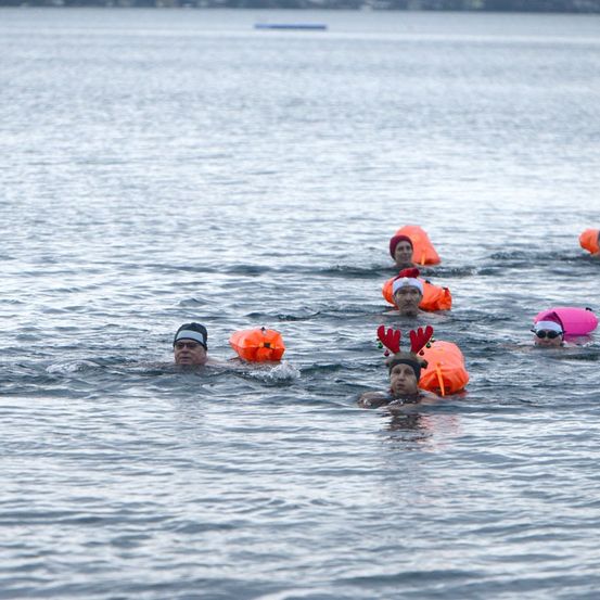 Eine Gruppe von Menschen schwimmt in einem See und trägt Schwimmwesten und festliche Mützen. Sie stehen in einer Reihe, und das Wasser spiegelt den Himmel wider.