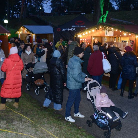 Ein nächtlicher Markt mit festlichen Lichtern, bei dem Menschen, darunter ein Mann, der einen Kinderwagen mit einem Kind schiebt, Stände erkunden. Die Atmosphäre ist warm mit Mänteln und Mützen.