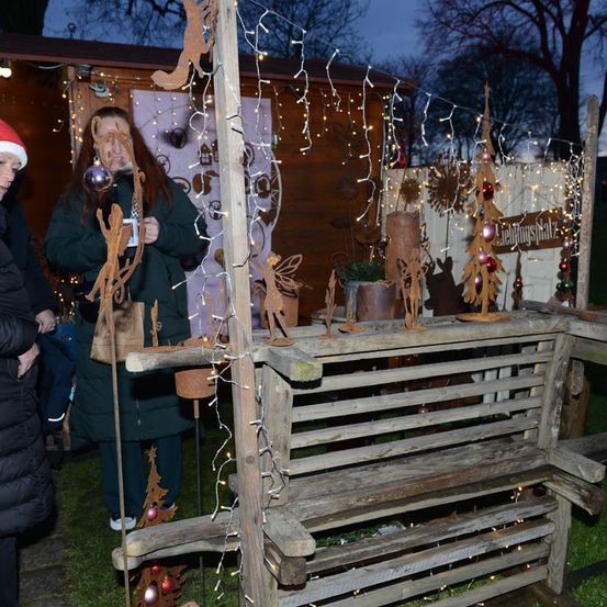 Eine Frau steht neben einer Holzbank, die mit Ornamenten und Elfenfiguren geschmückt ist, in einem Weihnachtsmarkt im Freien. Die Bank ist mit Lichterketten beleuchtet.
