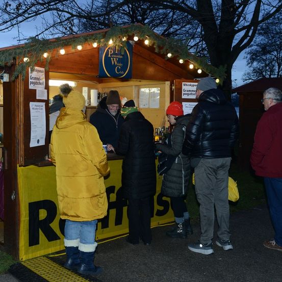 Menschen in Winterkleidung versammeln sich um einen Stand mit einem Banner, das 'Raffles' besagt, beleuchtet von Lichtern. Eine Person hinter dem Tresen bedient sie, mit Schildern für Gramnel und Whisky sichtbar.