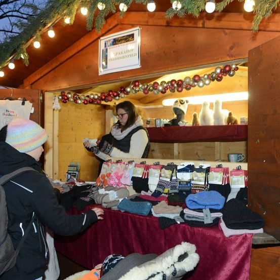 Eine Frau mit Brille steht in einem hölzernen Stand mit einem Schild, das 'Parahof Thersfeld' besagt. Eine Kundin mit Wintermütze stöbert durch einen Tisch voller Winterkleidung.