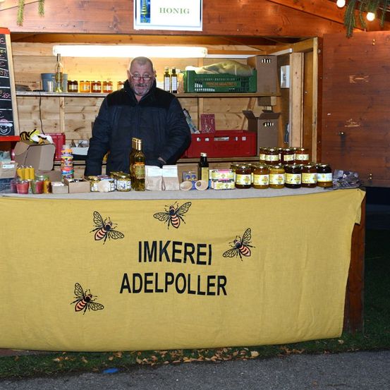 Ein Mann steht hinter einem gelben Tisch mit Honiggläsern und einem Schild, auf dem 'IMKEREI ADELPOLLER' steht. Dahinter befindet sich ein Holzregal mit verschiedenen Gläsern und Kisten.