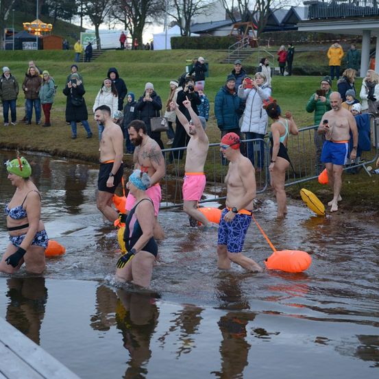 Eine Gruppe von Menschen geht in einem Fluss und hält Schwimmhilfen. Sie tragen Badeanzüge und Mützen. Einige von ihnen haben Tattoos. Am Ufer steht eine Menschenmenge und beobachtet. Einige halten Kameras und machen Fotos. Hinter der Menge befinden sich Bäume und ein Gebäude.