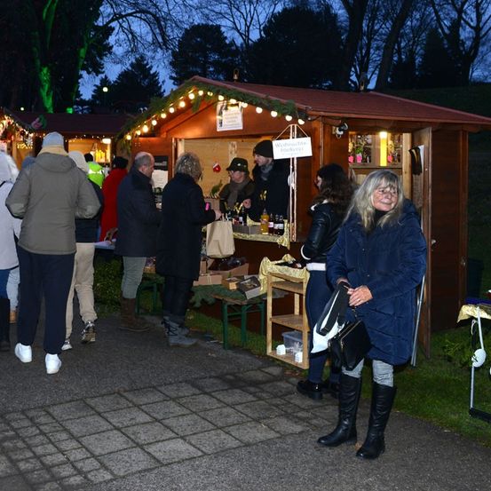Menschen versammeln sich bei Nacht um einen Essensstand, der von Lichterketten beleuchtet wird. Eine Frau lächelt und hält eine Handtasche. In der Nähe schenkt ein Mann in einem Hut Getränke ein.