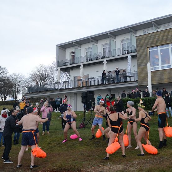 Eine Gruppe von Menschen, die in Badekleidung gekleidet sind und orangefarbene Luftballons halten, nehmen an einer Veranstaltung auf einem Rasenfeld vor einem Gebäude teil. Zuschauer beobachten von Balkonen und am Boden.