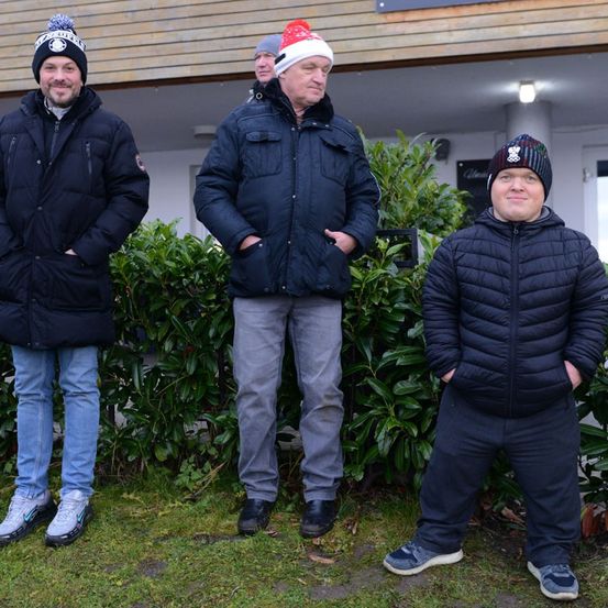 Vier Männer in Winterkleidung stehen vor einem Gebäude. Sie lächeln und scheinen für ein Foto zu posieren. Der Mann in der Mitte trägt eine Weihnachtsmütze.