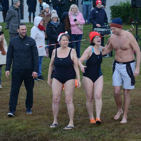 Eine Gruppe von Menschen steht auf einem Rasenfeld. Zwei Frauen in Badeanzügen gehen gemeinsam. Ein Mann steht neben ihnen. Dahinter lächelt ein Mann und schaut die beiden Frauen an. In der Ferne stehen Menschen und beobachten sie. Einige von ihnen tragen Mützen.