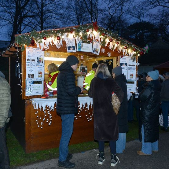 Eine Gruppe von Menschen steht in einer Schlange an einem Weihnachtsmarktstand, der mit Lichtern und Ornamenten geschmückt ist, während zwei Personen ihnen dienen.